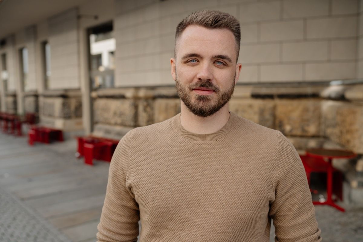 Max Krumbiegel im sandfarbenen Pullover vor einer Sandsteinmauer, er schaut ruhig und offen in die Kamera. Im Hintergrund unscharf die roten Stühle eines Cafés.