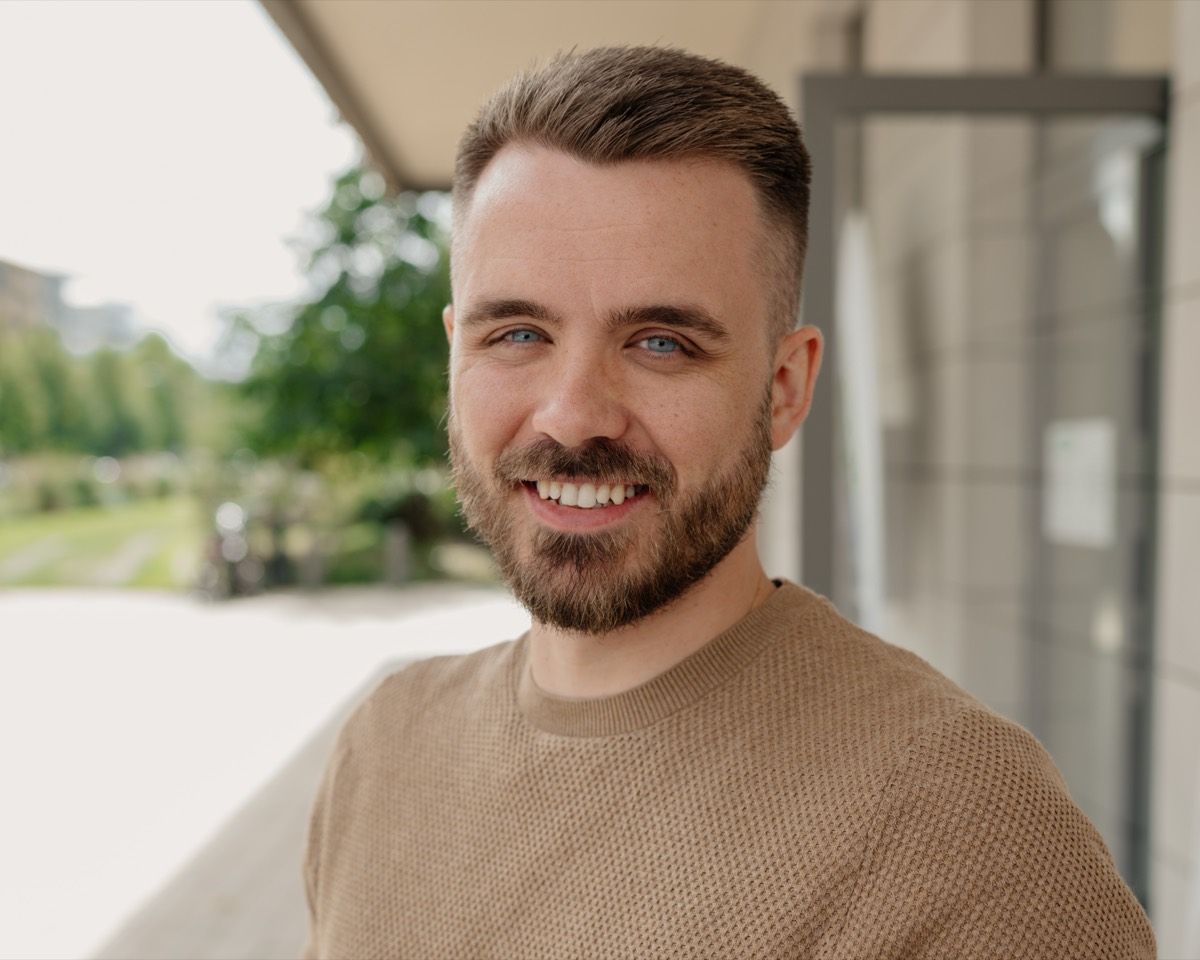 Max Krumbiegel lacht offen in die Kamera, nah aufgenommen. Hinter ihm unscharf ein Vordach, eine helle Fassade und grüne Bäume.